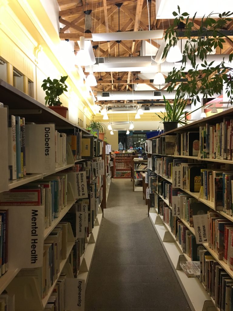 Image of a library aisle shows the very high ceilings, open rafters, and plants on tops of shelves