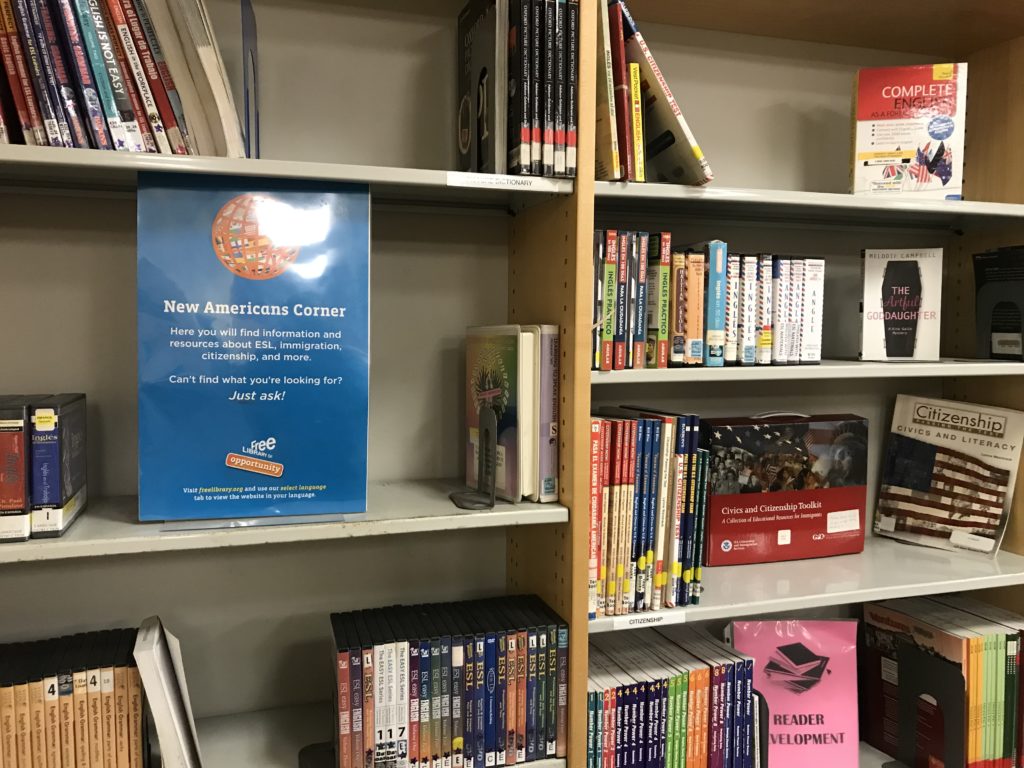 Bookshelves with paperback books, DVDs, books on CD, and a sign labeling the area as the New Americans Corner