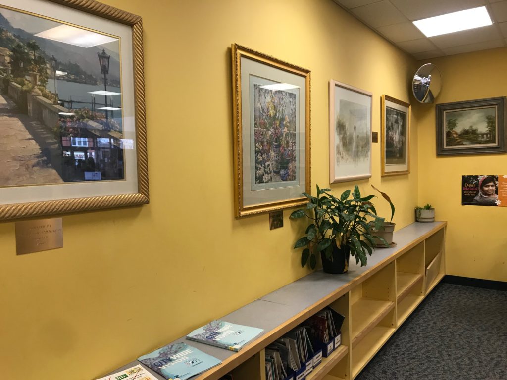 Low shelving with plants on top, and five framed works of art hanging on the walls