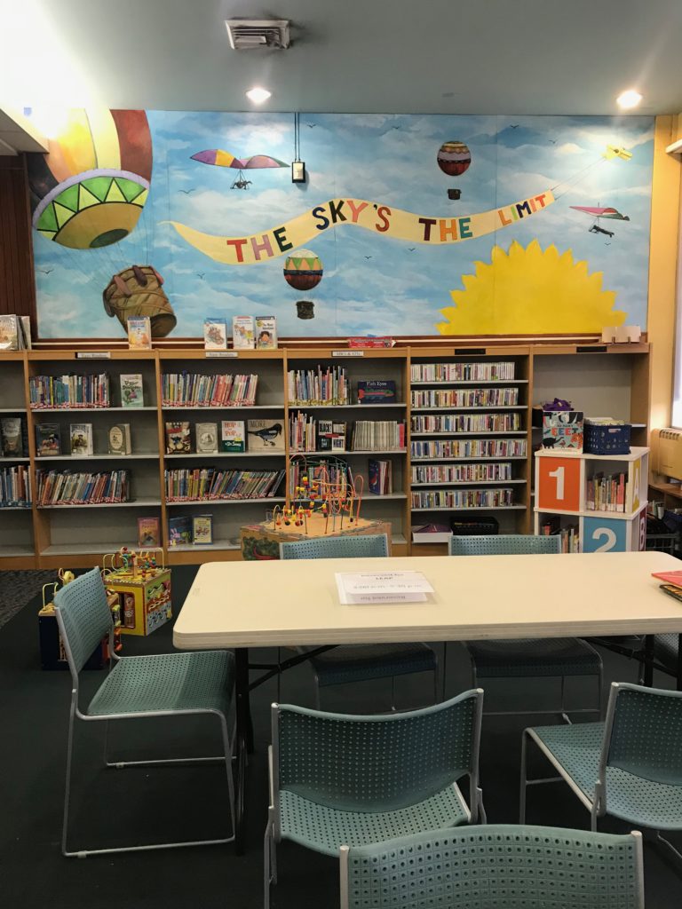 Table and chairs in front of bookshelves. Above the bookshelves, there is a brightly colored mural with a blue background and images of hot air balloons and the sun, plus the phrase "The sky's the limit."