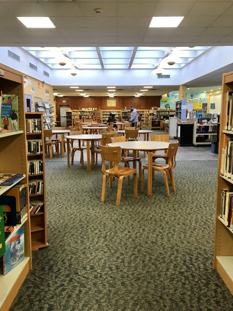 View into the middle of the library from between two shelves, with multiple sets of round tables and chairs throughout